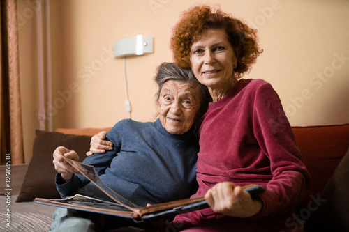 Senior woman and her adult daughter looking at photo album together on couch in living room, talking joyful discussing memories. Weekend with parents, family day, thanksgiving, mom's holiday
