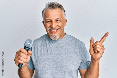 Middle age grey-haired man singing song using microphone smiling happy pointing with hand and finger to the side