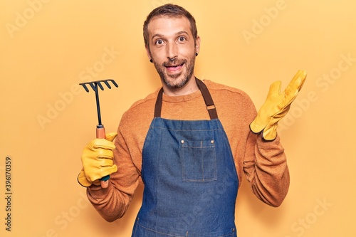 Young handsome man wearing gardener apron and gloves holding rake celebrating achievement with happy smile and winner expression with raised hand