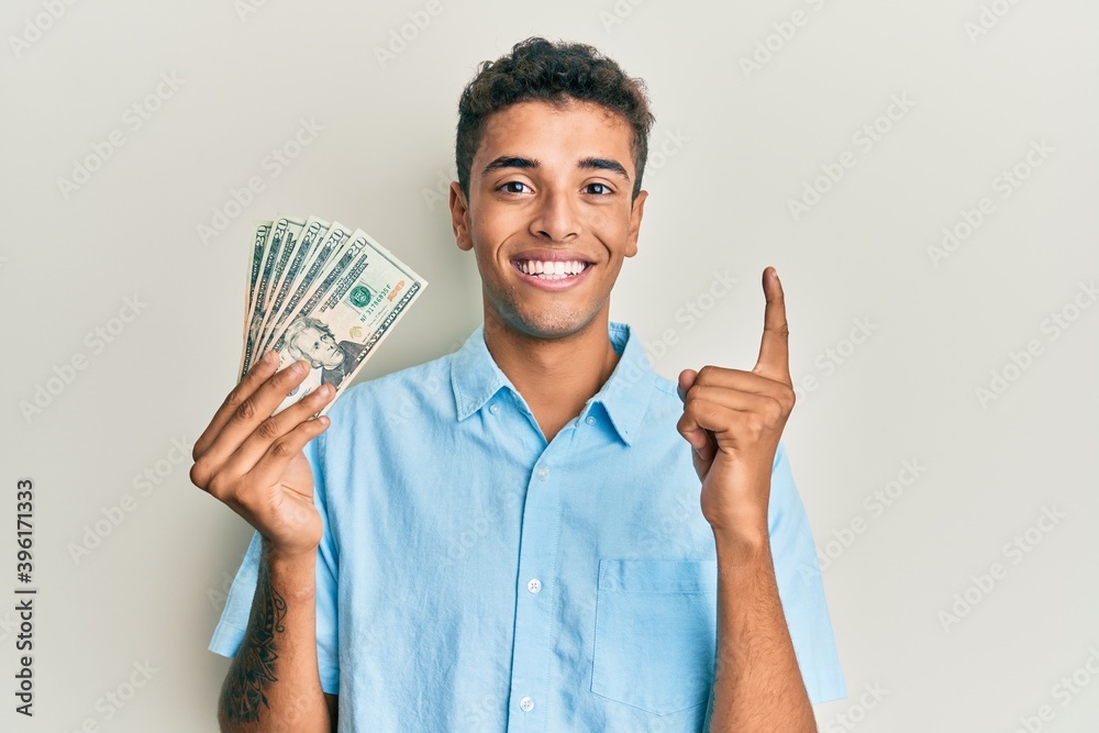 Young handsome african american man holding 20 dollars banknotes smiling with an idea or question pointing finger with happy face, number one Young handsome african american man holding 20 dollars banknotes smiling with an idea or question pointing finger with happy face, number one