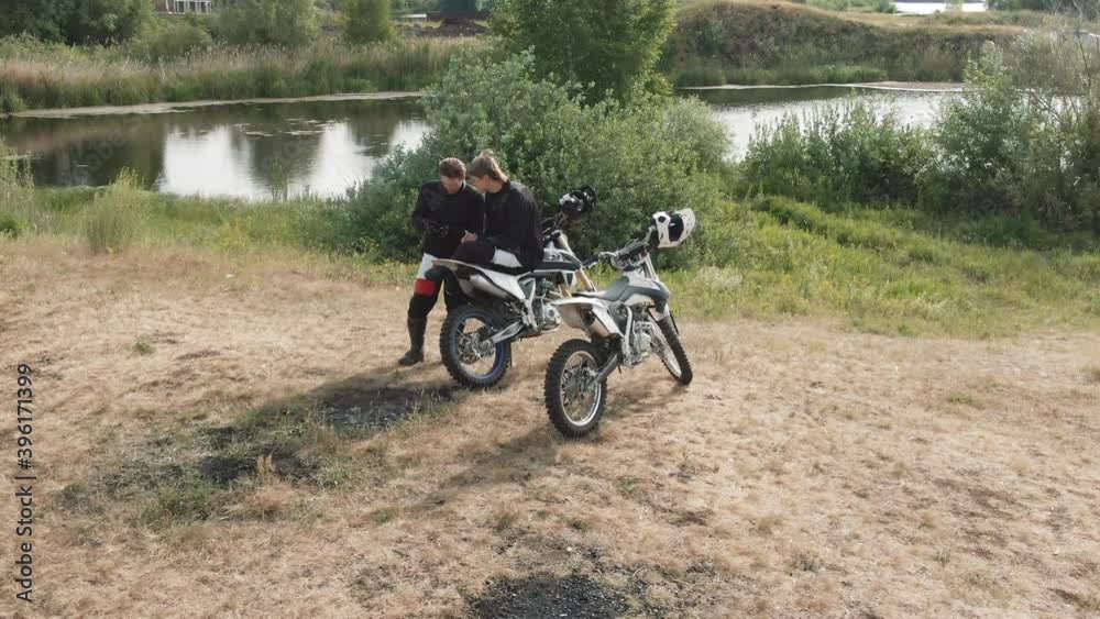 Drone shot of young male motorcyclists resting by lake and looking at mobile phone Drone shot of young male motorcyclists resting by lake and looking at mobile phone