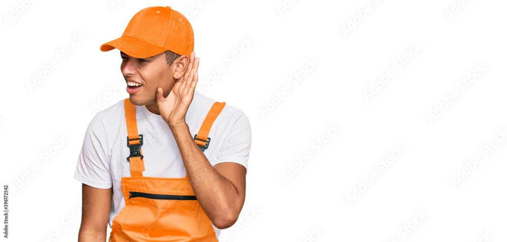 Young handsome african american man wearing handyman uniform smiling with hand over ear listening an hearing to rumor or gossip. deafness concept. Young handsome african american man wearing handyman uniform smiling with hand over ear listening an hearing to rumor or gossip. deafness concept.