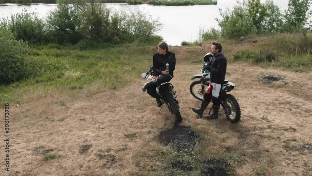 Tracking drone shot of two men with motorcycles chatting while drinking water and resting by lake Tracking drone shot of two men with motorcycles chatting while drinking water and resting by lake