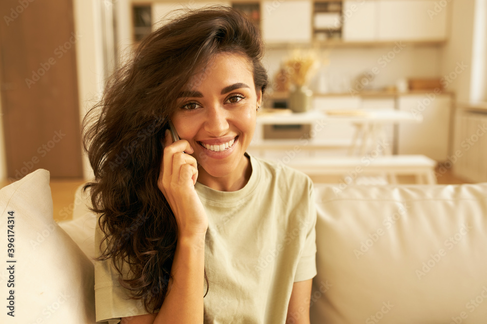 Gorgeous joyful young mixed race woman posing in stylish cozy home interior smiling happily while speaking on cell phone, sharing positive news. People, modern lifestyle and technology concept Gorgeous joyful young mixed race woman posing in stylish cozy home interior smiling happily while speaking on cell phone, sharing positive news. People, modern lifestyle and technology concept