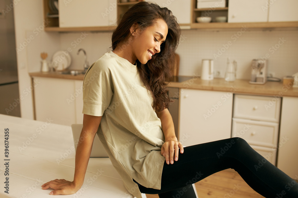 Side view of attractive young Hispanic female freelancer in comfy clothing sitting on dining table in cozy kitchen interior, relaxing while working from home. Pretty girl spending morning indoors Side view of attractive young Hispanic female freelancer in comfy clothing sitting on dining table in cozy kitchen interior, relaxing while working from home. Pretty girl spending morning indoors