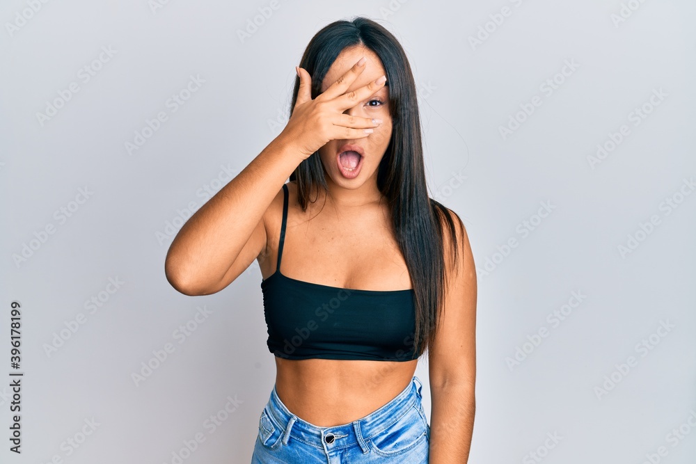 Young beautiful hispanic girl wearing casual clothes peeking in shock covering face and eyes with hand, looking through fingers afraid