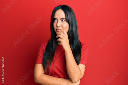 Young beautiful hispanic girl wearing casual red tshirt thinking concentrated about doubt with finger on chin and looking up wondering