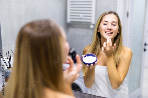 beauty, skin care and people concept. Young woman applying cream to face and looking to mirror at home bathroom