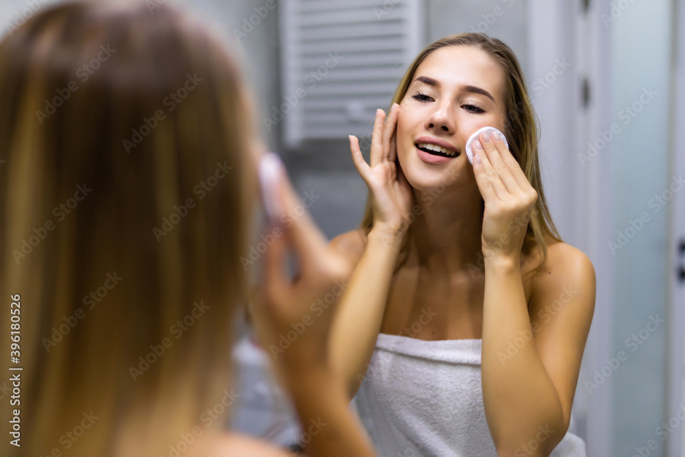 Beauty, skin care and people concept - smiling young woman applying lotion to cotton disc for washing her face at bathroom Beauty, skin care and people concept - smiling young woman applying lotion to cotton disc for washing her face at bathroom