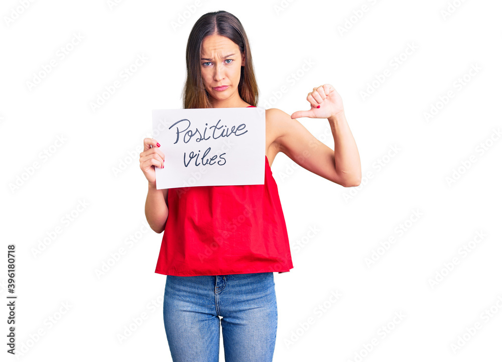 Beautiful brunette young woman holding positive vibes banner with angry face, negative sign showing dislike with thumbs down, rejection concept Beautiful brunette young woman holding positive vibes banner with angry face, negative sign showing dislike with thumbs down, rejection concept