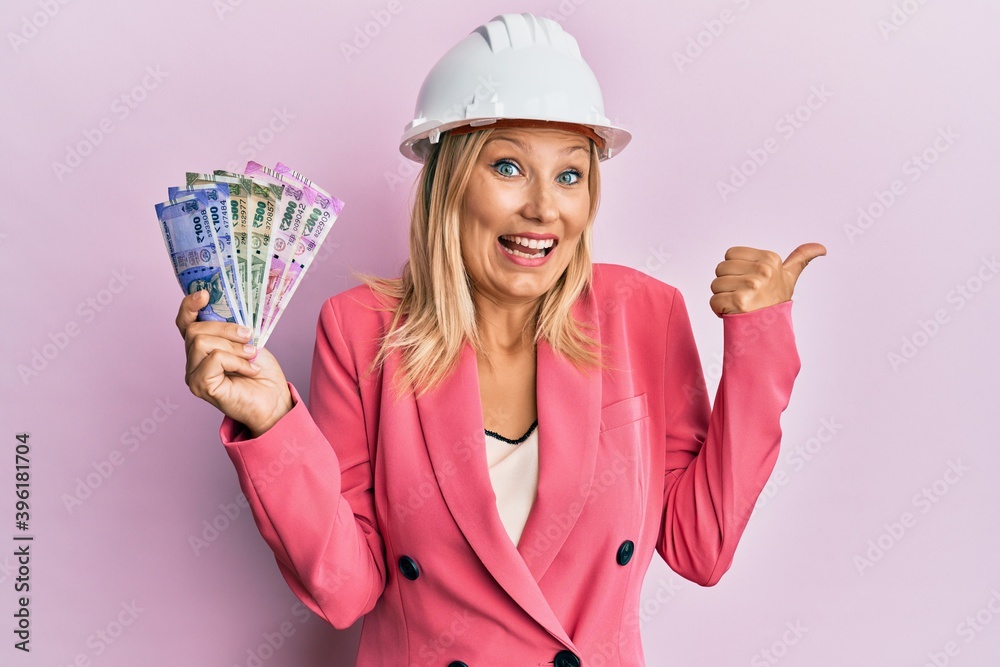 Beautiful middle age blonde woman wearing architect hardhat holding indian rupee pointing thumb up to the side smiling happy with open mouth Beautiful middle age blonde woman wearing architect hardhat holding indian rupee pointing thumb up to the side smiling happy with open mouth