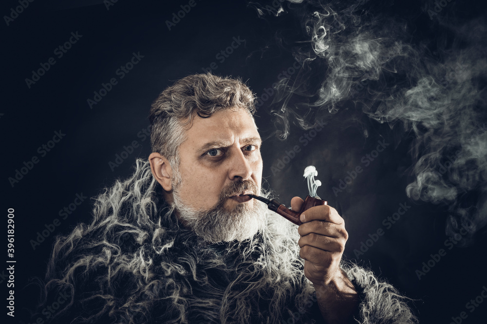 Fototapeta premium Solid bearded man in a fur mantle smoking pipe