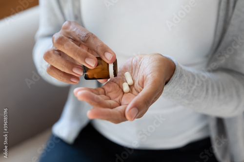 Crop close up of African American woman take pills or tablets from bottle. Biracial female have nutritional dietary supplements or medicines from packaging. Healthcare, pharmacy, wellness concept.