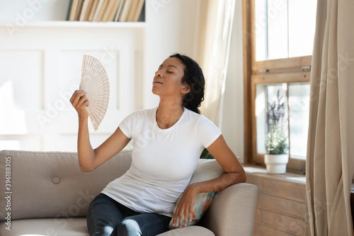 Overheated millennial African American woman sit relax on sofa at home breathe fresh air from waver. Unwell young biracial female renter suffer from heatstroke hot weather wave with hand fan.