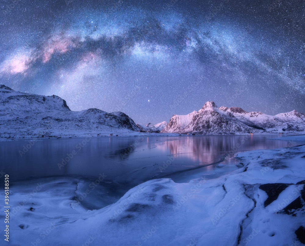 Milky Way above frozen sea coast and snow covered mountains in winter at night in Lofoten Islands, Norway. Arctic landscape with blue starry sky, water, ice, snowy rocks, milky way. Space and galaxy