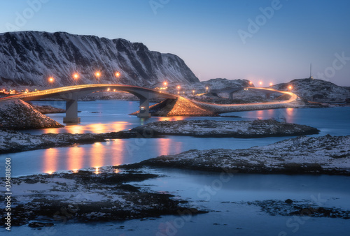 Bridge with illumination, snow covered mountains, village and blue sky with reflection in water. Night landscape with bridge, snowy rocks, city lights, sea. Winter in Lofoten islands, Norway. Road © den-belitsky