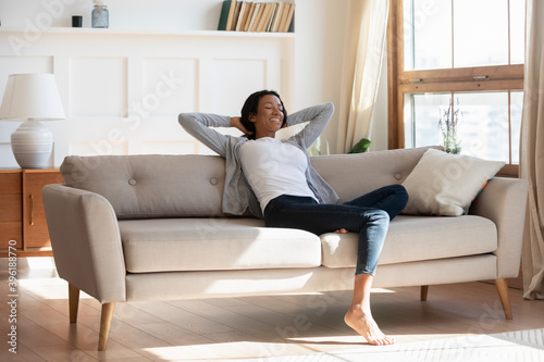 Happy young African American young woman relax on couch in living room relieve negative emotions. Smiling calm biracial female tenant rest on sofa at home, breathe fresh air. Stress free concept.