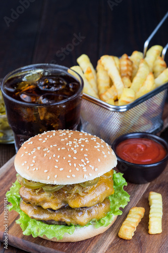 Double cheese burger with two beef patty, cheddar cheese, lettuce, caramelized onion and jalapeno slices, served with french fries and soda, on wooden board, vertical