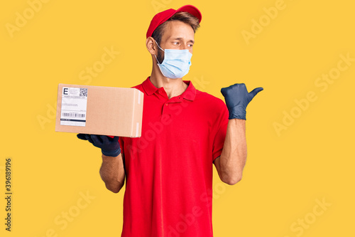 Handsome blond man with beard holding delivery box wearing medical mask pointing thumb up to the side smiling happy with open mouth