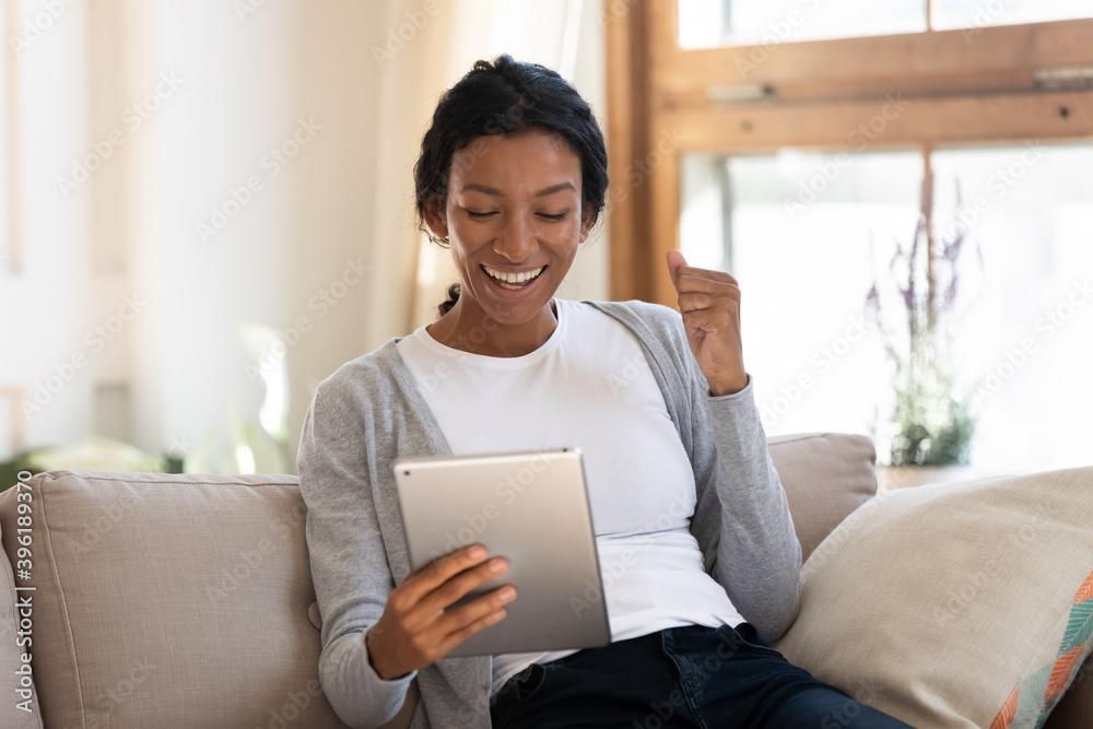 Excited millennial biracial woman relax on couch at home triumph win lottery online on tablet. Happy overjoyed young African American female look at pad screen feel euphoric with good message. Excited millennial biracial woman relax on couch at home triumph win lottery online on tablet. Happy overjoyed young African American female look at pad screen feel euphoric with good message.