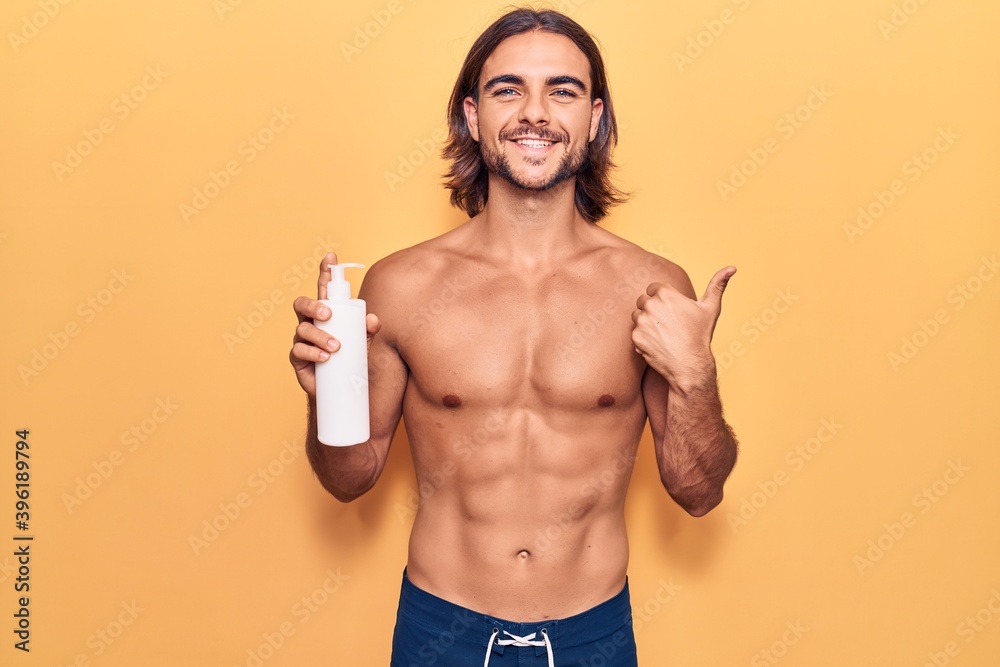 Young handsome man wearing swimwear holding sunscreen lotion smiling happy and positive, thumb up doing excellent and approval sign