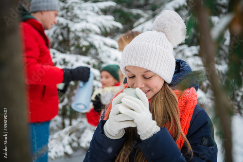 A happy teenage girl enjoys the first snow and drinks hot tea from a mug in the winter forest during the Christmas holidays