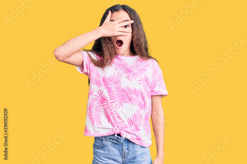 Cute hispanic child girl wearing casual clothes peeking in shock covering face and eyes with hand, looking through fingers with embarrassed expression.