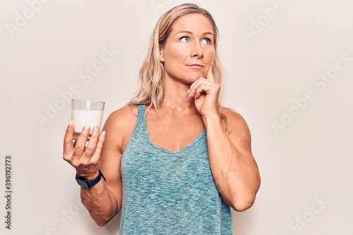 Middle age caucasian blonde woman holding a glass of milk serious face thinking about question with hand on chin, thoughtful about confusing idea