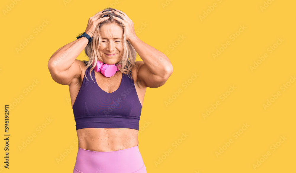 Middle age fit blonde woman wearing gym clothes and using headphones suffering from headache desperate and stressed because pain and migraine. hands on head. Middle age fit blonde woman wearing gym clothes and using headphones suffering from headache desperate and stressed because pain and migraine. hands on head.