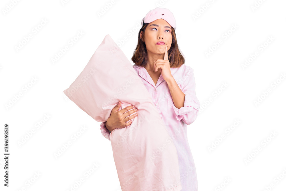 Young beautiful chinese girl wearing sleep mask and pajama holding pillow serious face thinking about question with hand on chin, thoughtful about confusing idea Young beautiful chinese girl wearing sleep mask and pajama holding pillow serious face thinking about question with hand on chin, thoughtful about confusing idea