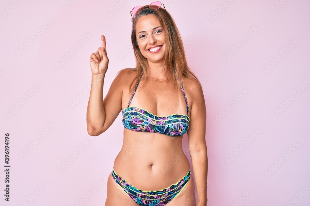 Middle age hispanic woman wearing bikini showing and pointing up with finger number one while smiling confident and happy. Middle age hispanic woman wearing bikini showing and pointing up with finger number one while smiling confident and happy.