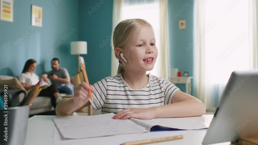 Small beautiful blonde girl is sitting at work desk at home, has online lesson, raising hand and talking to her teacher, use tablet computer, parents are sitting on sofa, Foreground, Slow motion. Small beautiful blonde girl is sitting at work desk at home, has online lesson, raising hand and talking to her teacher, use tablet computer, parents are sitting on sofa, Foreground, Slow motion.