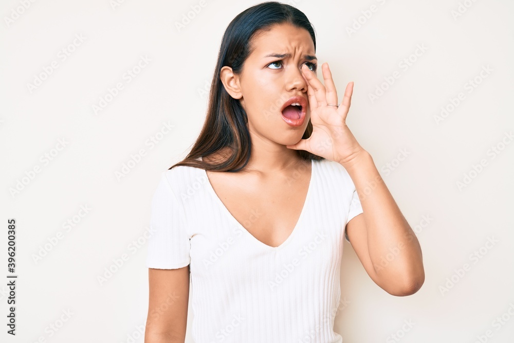 Young beautiful latin girl wearing casual white tshirt shouting and screaming loud to side with hand on mouth. communication concept. Young beautiful latin girl wearing casual white tshirt shouting and screaming loud to side with hand on mouth. communication concept.
