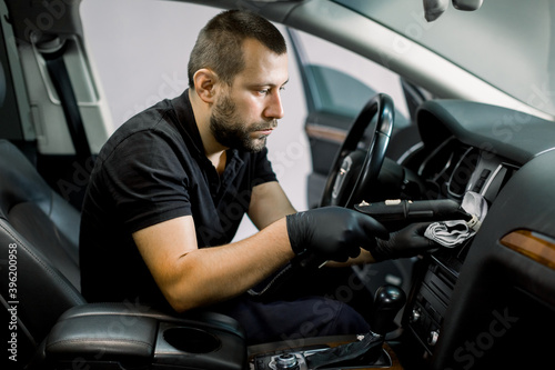 Auto cleaning service and detailing concept. Handsome Caucasian man in black uniform and rubber gloves, cleaning interior of the car with hot steam cleaner. Selective focus.