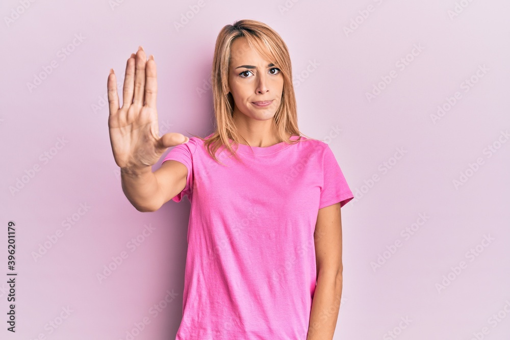 Beautiful blonde woman wearing casual pink tshirt doing stop sing with palm of the hand. warning expression with negative and serious gesture on the face. Beautiful blonde woman wearing casual pink tshirt doing stop sing with palm of the hand. warning expression with negative and serious gesture on the face.