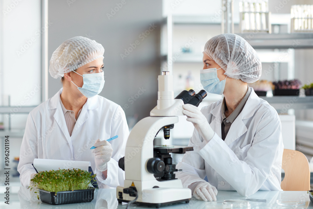 Portrait of two young female scientists looking in microscope while studying plant samples in biotechnology lab, copy space Portrait of two young female scientists looking in microscope while studying plant samples in biotechnology lab, copy space