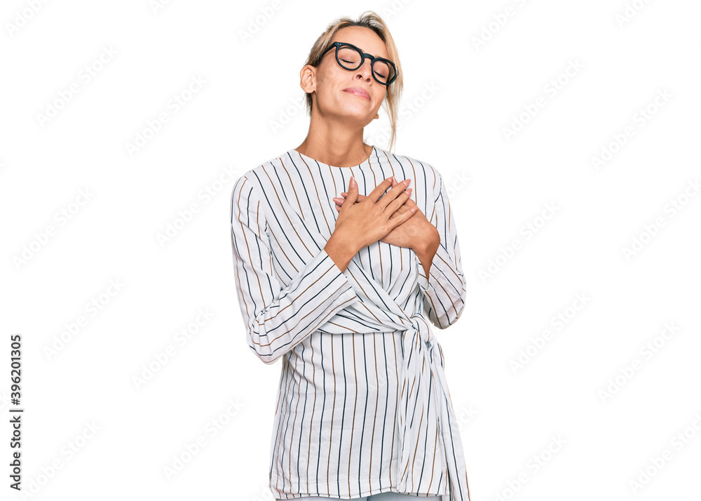 Beautiful blonde woman wearing business shirt and glasses smiling with hands on chest with closed eyes and grateful gesture on face. health concept. Beautiful blonde woman wearing business shirt and glasses smiling with hands on chest with closed eyes and grateful gesture on face. health concept.