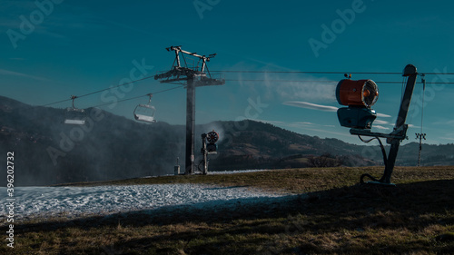 View of snow cannons or artificial snow makers on a ski slope on a sunny day. Visible partly covered ski slope with snow and some grass too