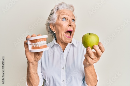 Senior grey-haired woman holding green apple and denture teeth angry and mad screaming frustrated and furious, shouting with anger looking up.
