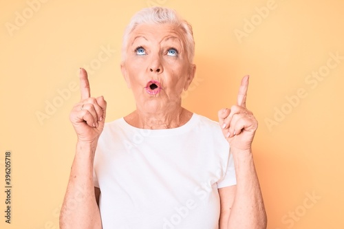 Senior beautiful woman with blue eyes and grey hair wearing classic white tshirt over yellow background amazed and surprised looking up and pointing with fingers and raised arms.