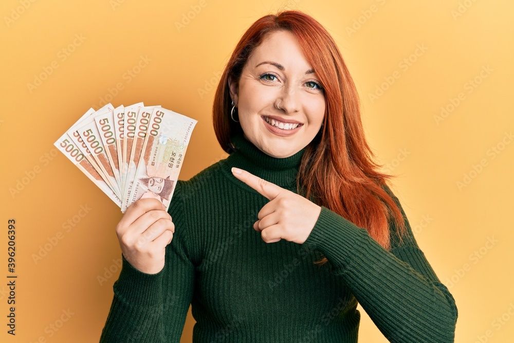 Beautiful redhead woman holding 5000 south korean won banknotes smiling happy pointing with hand and finger