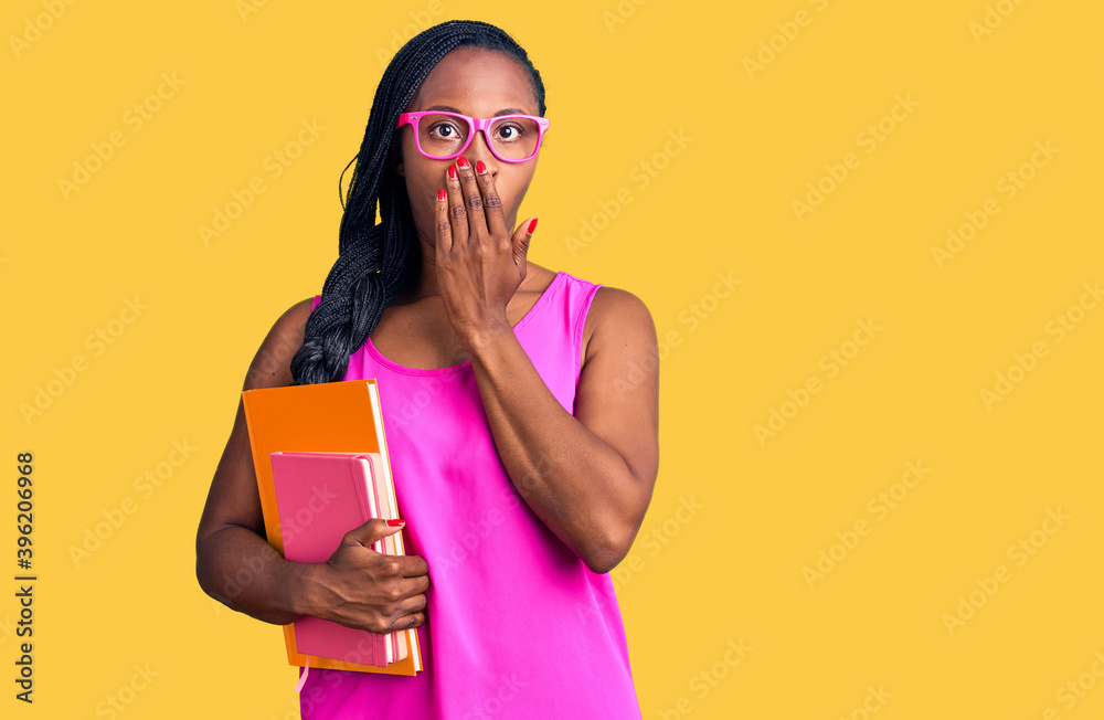 Young african american woman wearing glasses holding book covering mouth with hand, shocked and afraid for mistake. surprised expression Young african american woman wearing glasses holding book covering mouth with hand, shocked and afraid for mistake. surprised expression