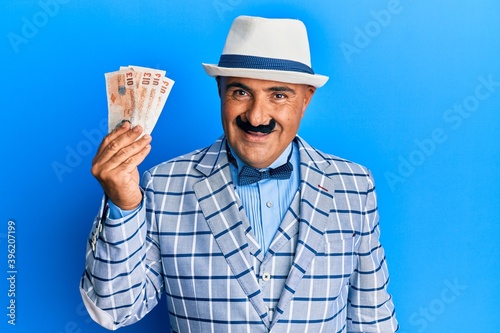 Mature middle east man with mustache wearing elegant vintage style holding pounds banknotes looking positive and happy standing and smiling with a confident smile showing teeth photo