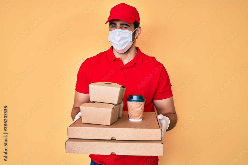 Handsome hispanic man with beard holding delivery food wearing medical mask winking looking at the camera with sexy expression, cheerful and happy face. Handsome hispanic man with beard holding delivery food wearing medical mask winking looking at the camera with sexy expression, cheerful and happy face.