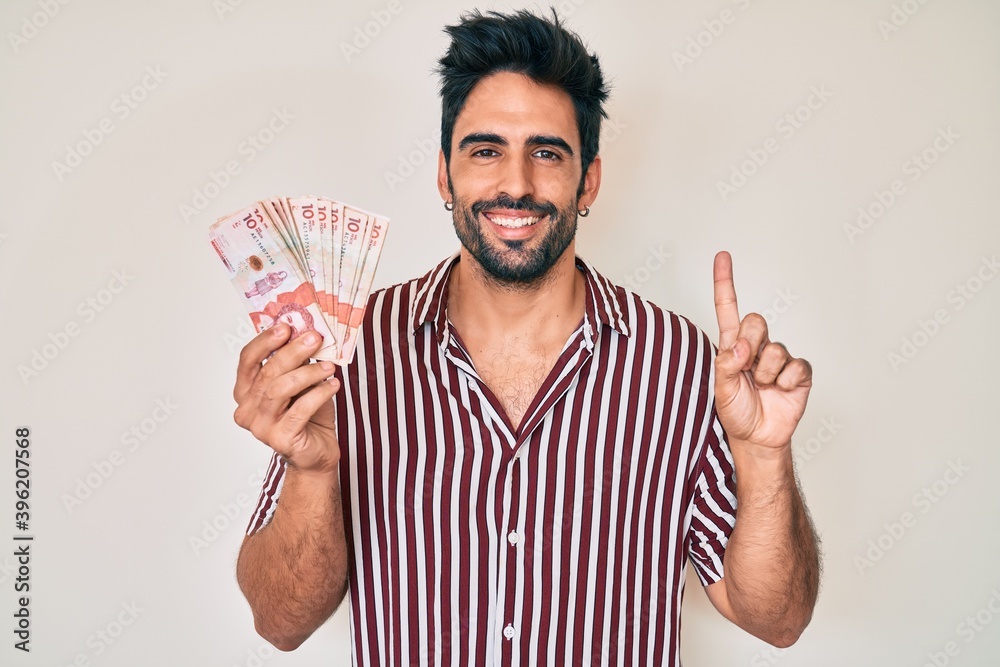 Handsome hispanic man with beard holding colombian pesos smiling with an idea or question pointing finger with happy face, number one Handsome hispanic man with beard holding colombian pesos smiling with an idea or question pointing finger with happy face, number one