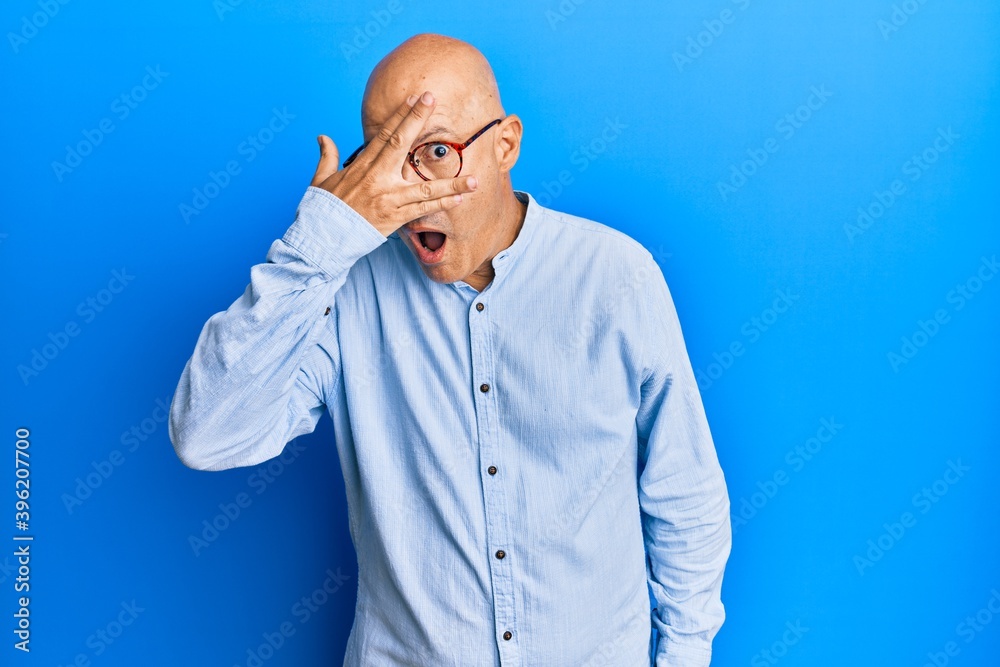Middle age bald man wearing casual clothes and glasses peeking in shock covering face and eyes with hand, looking through fingers with embarrassed expression. Middle age bald man wearing casual clothes and glasses peeking in shock covering face and eyes with hand, looking through fingers with embarrassed expression.