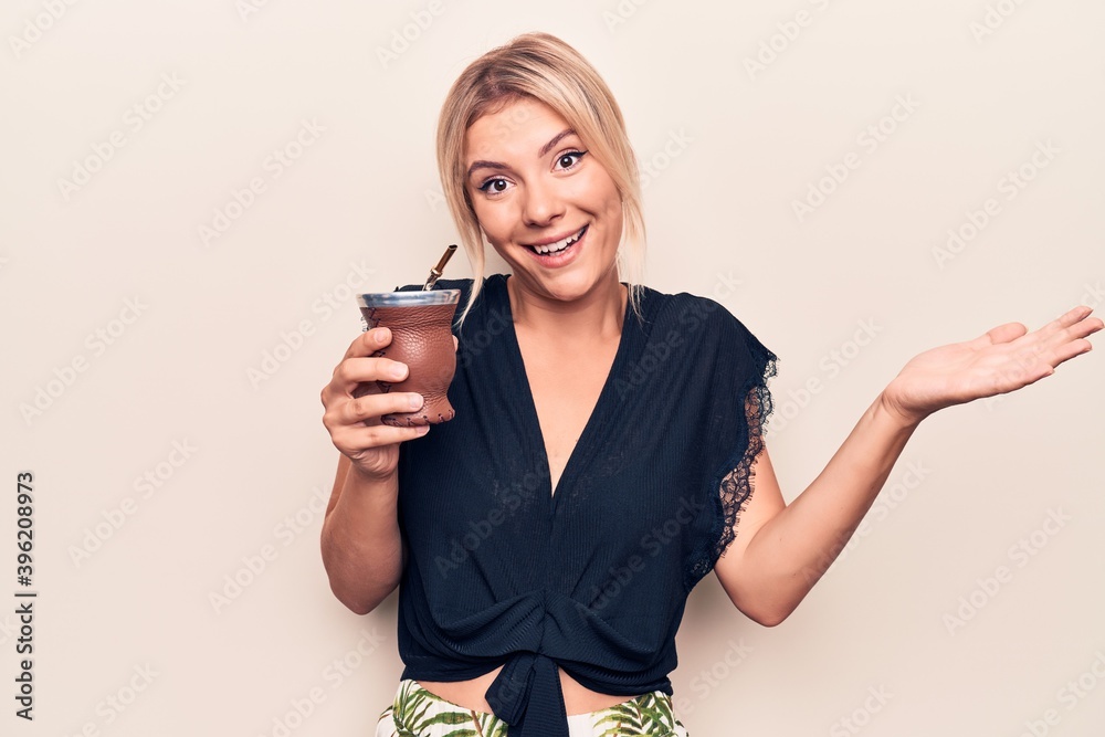 Young beautiful blonde woman drinking cup of mate infusion beverage over white background celebrating achievement with happy smile and winner expression with raised hand Young beautiful blonde woman drinking cup of mate infusion beverage over white background celebrating achievement with happy smile and winner expression with raised hand
