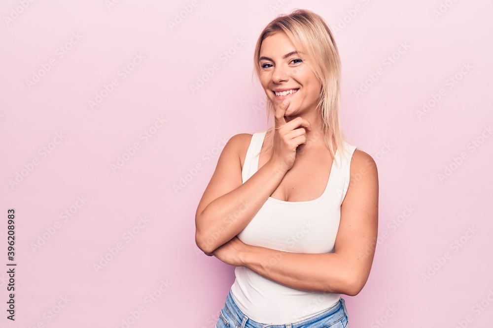 Young beautiful blonde woman wearing casual t-shirt standing over isolated pink background smiling looking confident at the camera with crossed arms and hand on chin. Thinking positive. Young beautiful blonde woman wearing casual t-shirt standing over isolated pink background smiling looking confident at the camera with crossed arms and hand on chin. Thinking positive.