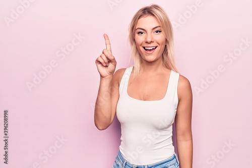 Young beautiful blonde woman wearing casual t-shirt standing over isolated pink background smiling with an idea or question pointing finger up with happy face, number one
