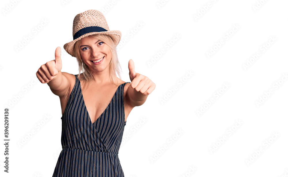 Young beautiful blonde woman wearing summer hat and dress approving doing positive gesture with hand, thumbs up smiling and happy for success. winner gesture. Young beautiful blonde woman wearing summer hat and dress approving doing positive gesture with hand, thumbs up smiling and happy for success. winner gesture.